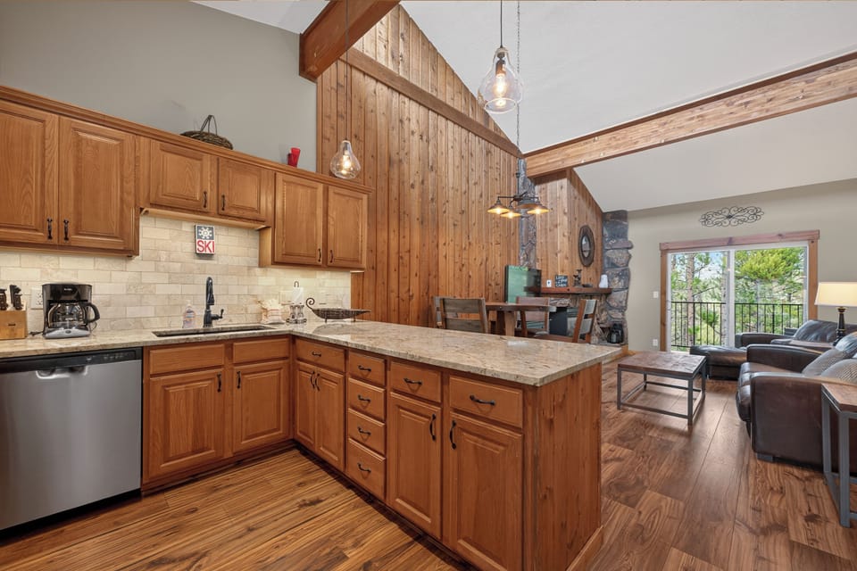 Relax in your open-concept space featuring a fully equipped kitchen with granite countertops, vaulted ceilings with rustic wood accents, and a cozy stone fireplace perfect for mountain getaways.