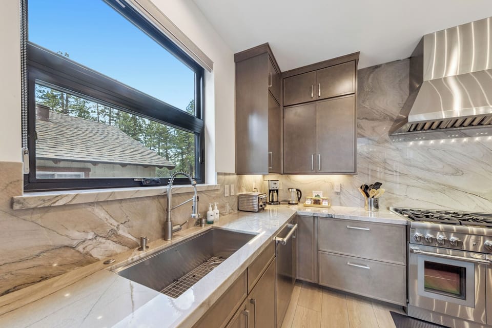 Picture yourself preparing meals in this sleek, modern kitchen with gleaming marble countertops, professional-grade stainless appliances, and natural light streaming through expansive windows overlooking peaceful trees.