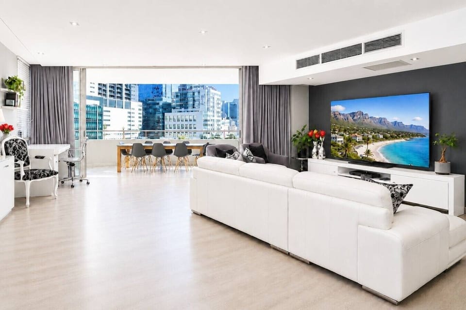 Bright and spacious open-plan living area designed for comfort and entertaining, featuring stylish furnishings, a large smart TV, dedicated dining space, and expansive city views through oversized windows.