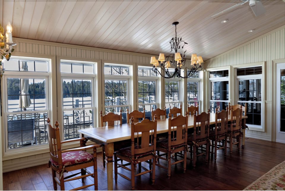 Dining room with long wooden table, fourteen high-back chairs with red cushions, and large windows overlooking the lake.