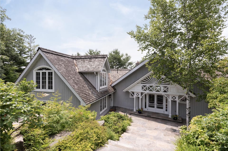 Gray cottage exterior with white-trimmed windows, decorative porch details, stone steps, and lush green landscaping.