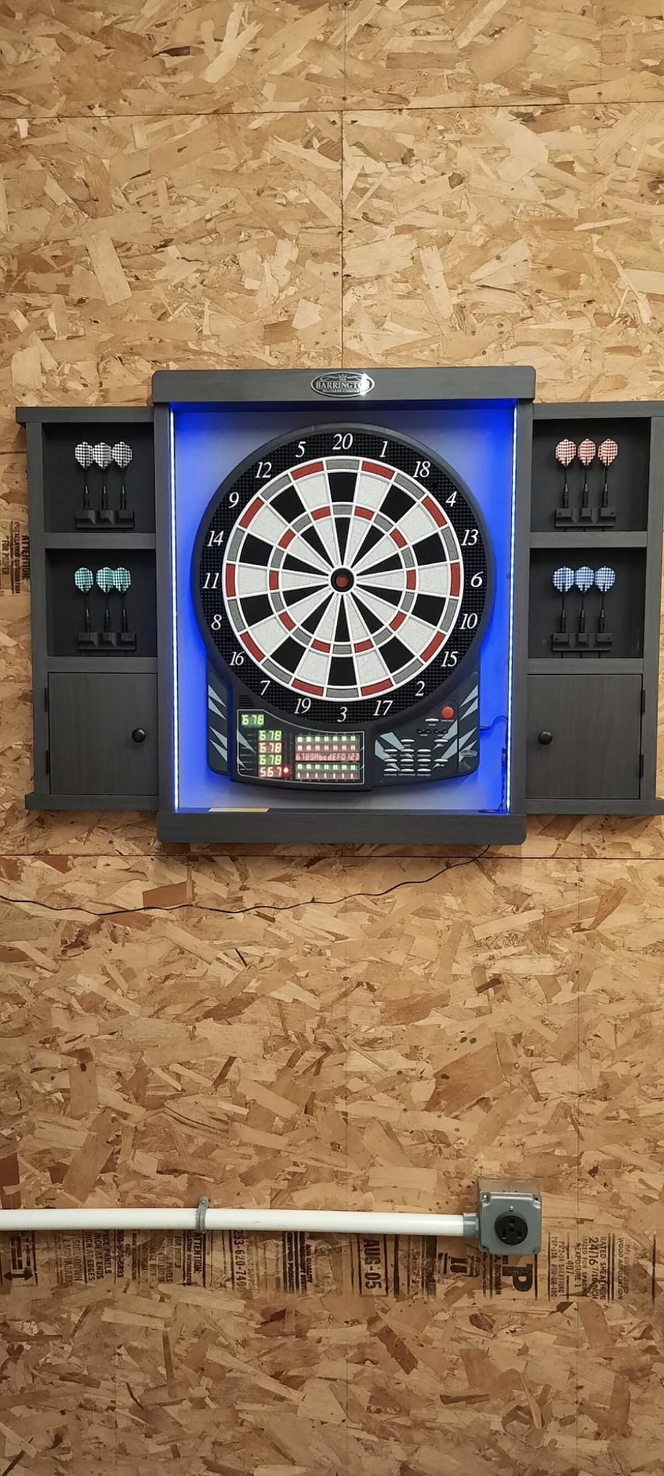 Electronic dartboard with blue LED backlight mounted on textured plywood wall, flanked by six sets of darts.