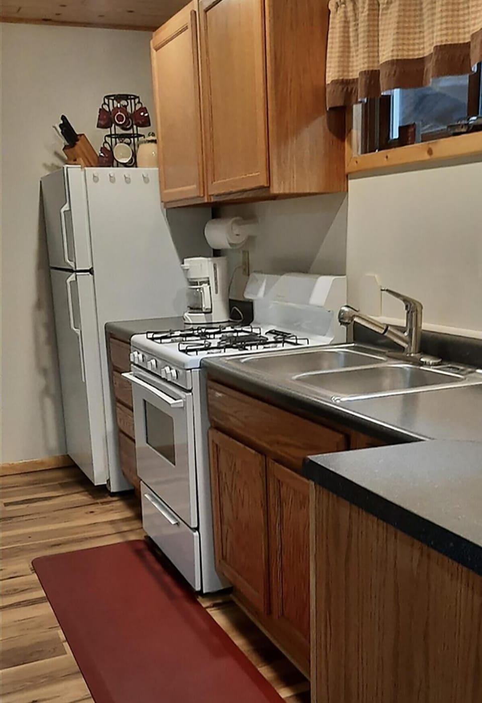 Kitchen features oak cabinets, white gas stove, double stainless sink, and refrigerator on wood plank flooring.