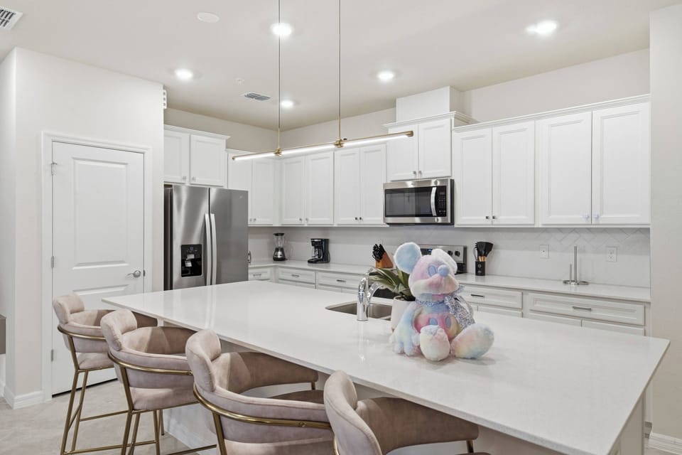 Cook and gather in a bright modern kitchen with white cabinets, stainless steel appliances, a spacious island, soft pendant lighting, plush bar chairs, and playful Mickey decor, perfect for family meals during vacation.