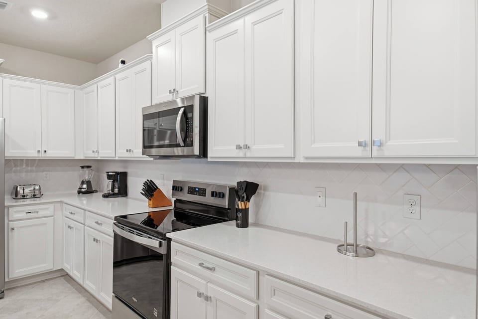 Cook with ease in a bright modern kitchen with white cabinets, stainless steel appliances, spacious counters, a large island, soft lighting, and an open view to the living room, perfect for family meals during vacation.