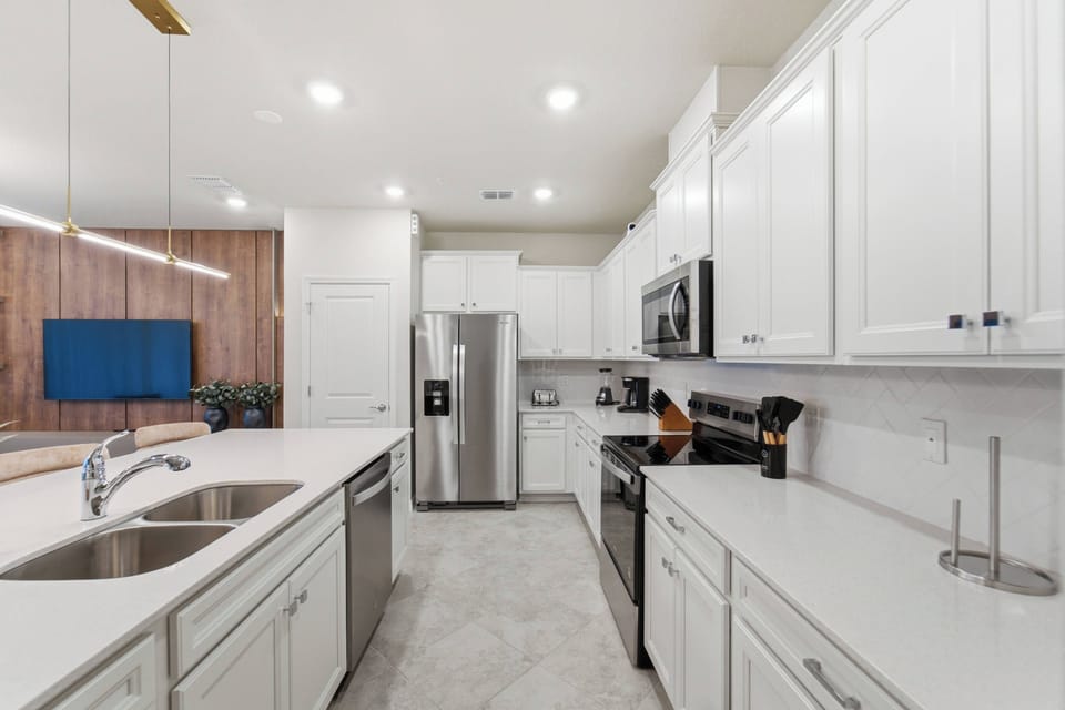 Cook with ease in a bright modern kitchen with white cabinets, stainless steel appliances, spacious counters, a large island, soft lighting, and an open view to the living room, perfect for family meals during vacation.