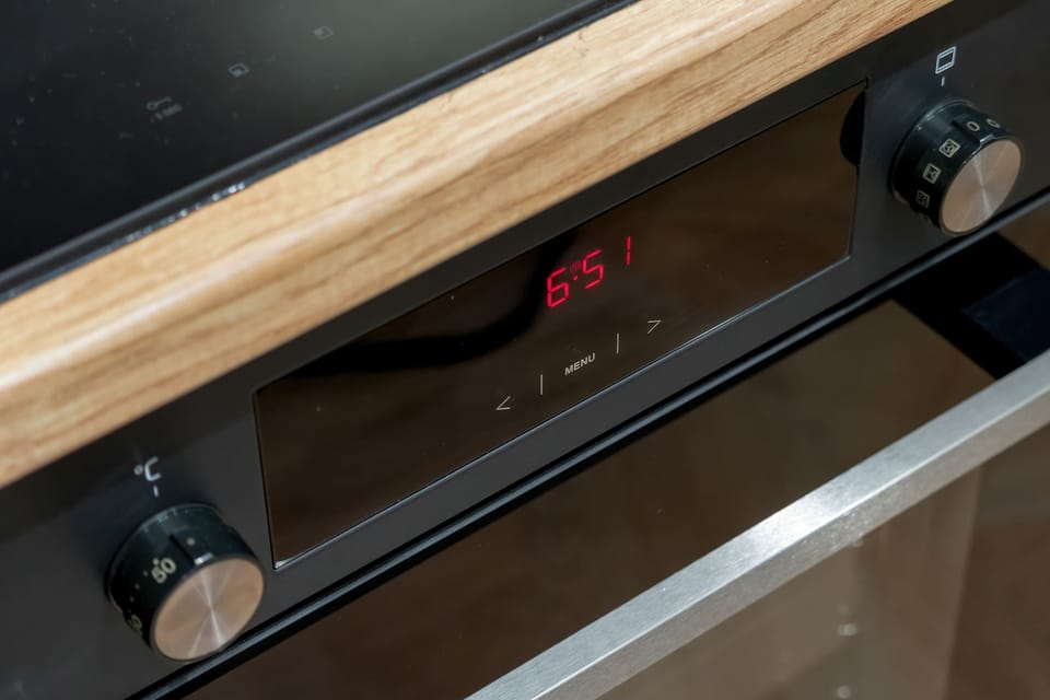 A close-up shot focuses on the digital display and control knobs of a modern black oven. The appliance is built into a wooden countertop framework.