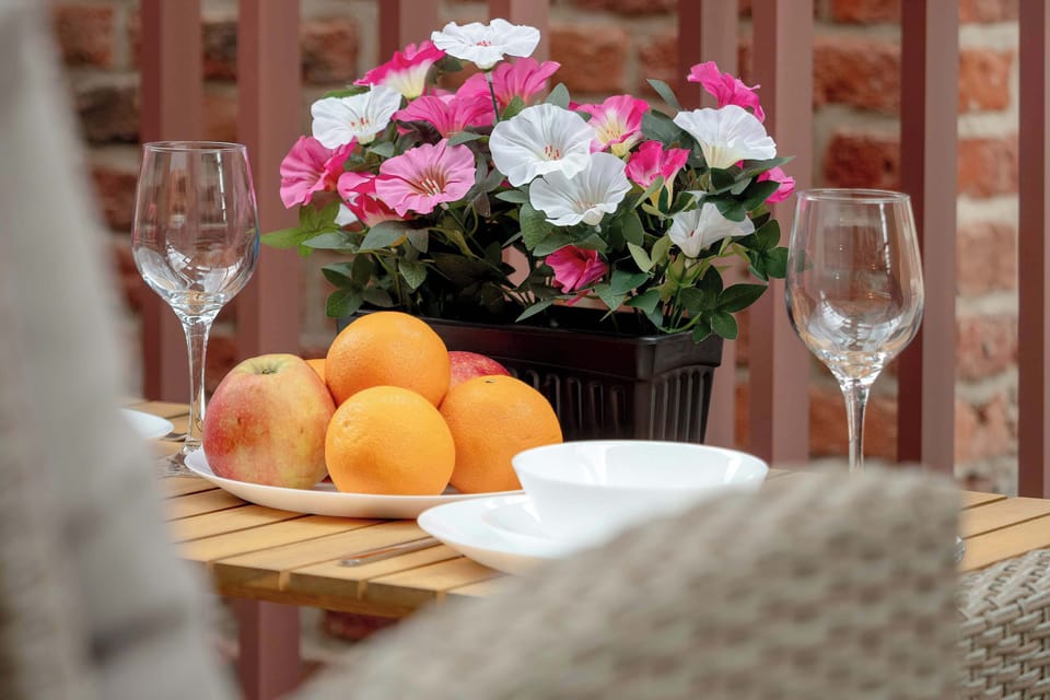A wooden table on the terrace is set with wine glasses and a plate filled with fresh oranges and apricots. A vibrant planter box with pink and white flowers sits beautifully in the background.