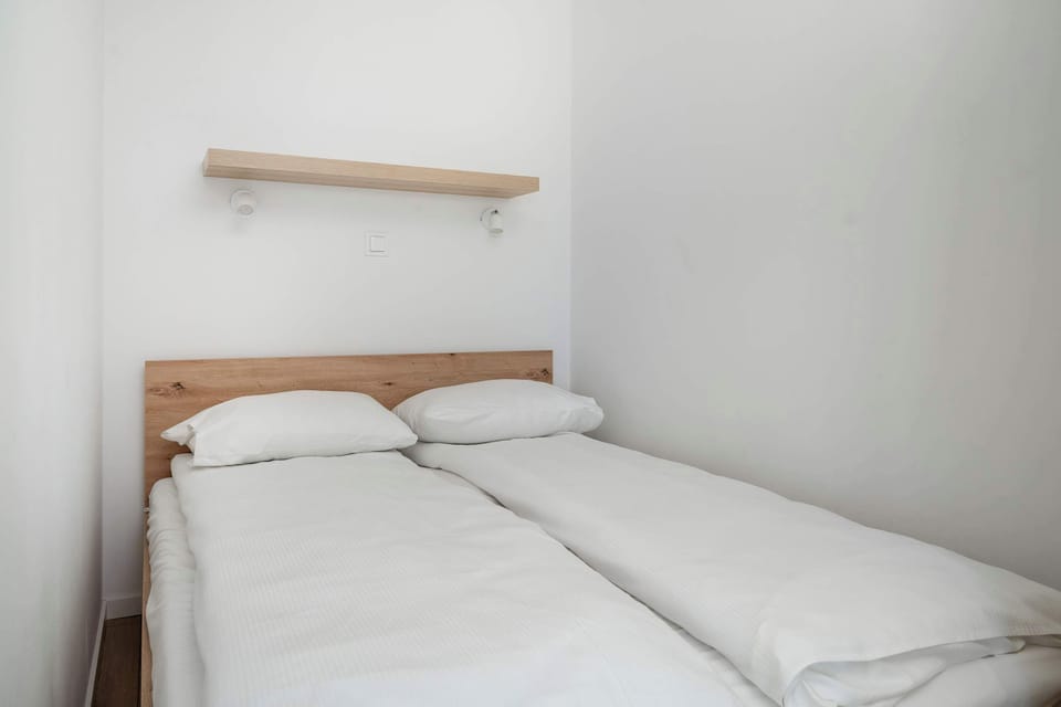 A minimalist bedroom features a double bed neatly made with crisp white linens tucked against white walls. A simple light wood shelf is mounted on the wall directly above the head of the bed.