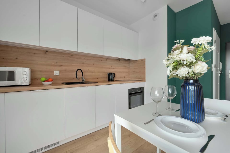 A sleek kitchen setup displays minimalist white cabinets contrasted by a warm, light wood countertop and backsplash. A dining table decorated with flowers and glassware stands in the foreground.