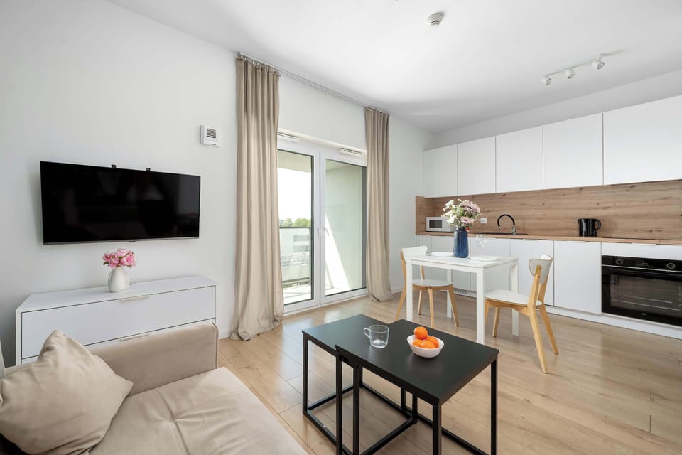A spacious studio layout reveals a wall-mounted television across from a beige sofa and black coffee table. The white kitchen units and a dining area are clearly visible in the background.