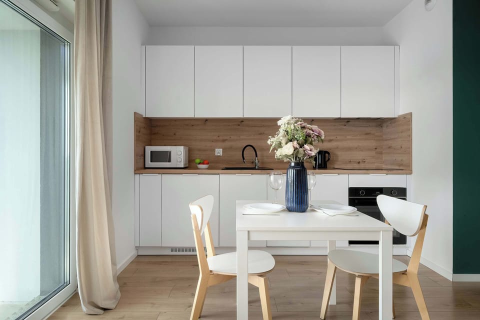 A bright, open-plan kitchen and dining area features white upper cabinets and a light wood backsplash and countertop. A white dining table set for two sits in the foreground against a dark green wall backdrop.