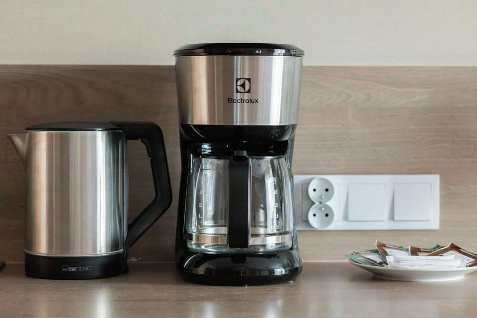 A close-up detail of the kitchen counter showcasing a stainless steel electric kettle and a filter coffee machine. A small plate with a treat sits next to them against the light wood backdrop.
