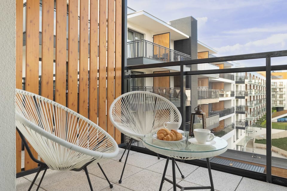 A private balcony area showing two modern, white wicker chairs and a small glass-topped table. The glass railing overlooks other modern residential or holiday apartment buildings.