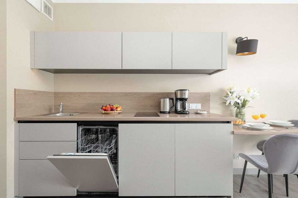 The same minimalist kitchen counter layout, but with one of the lower dishwasher doors left open. A coffee maker, juice, and a bowl of fresh fruit are neatly arranged on the surface.