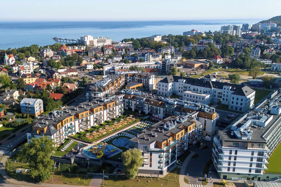 An aerial drone shot showcasing a large, modern residential complex situated near a coastline. The buildings feature dark roofs and light facades, surrounded by lush green trees and a body of water in the distance.