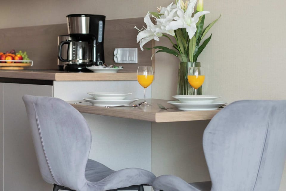 A cozy dining nook featuring a small wooden table set against the kitchen counter with two plush gray chairs. Two glasses of orange juice and a vase of white lilies create an inviting atmosphere.