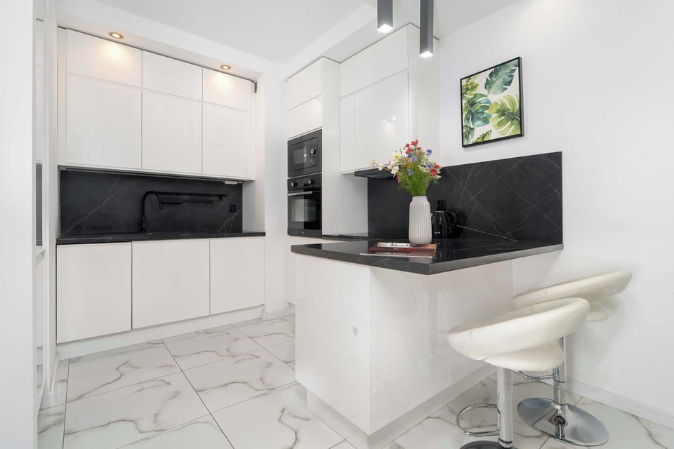 The bright, open-concept kitchenette showcases clean white cabinetry, dark contrasting countertops, and a small breakfast bar with a stylish bar stool.