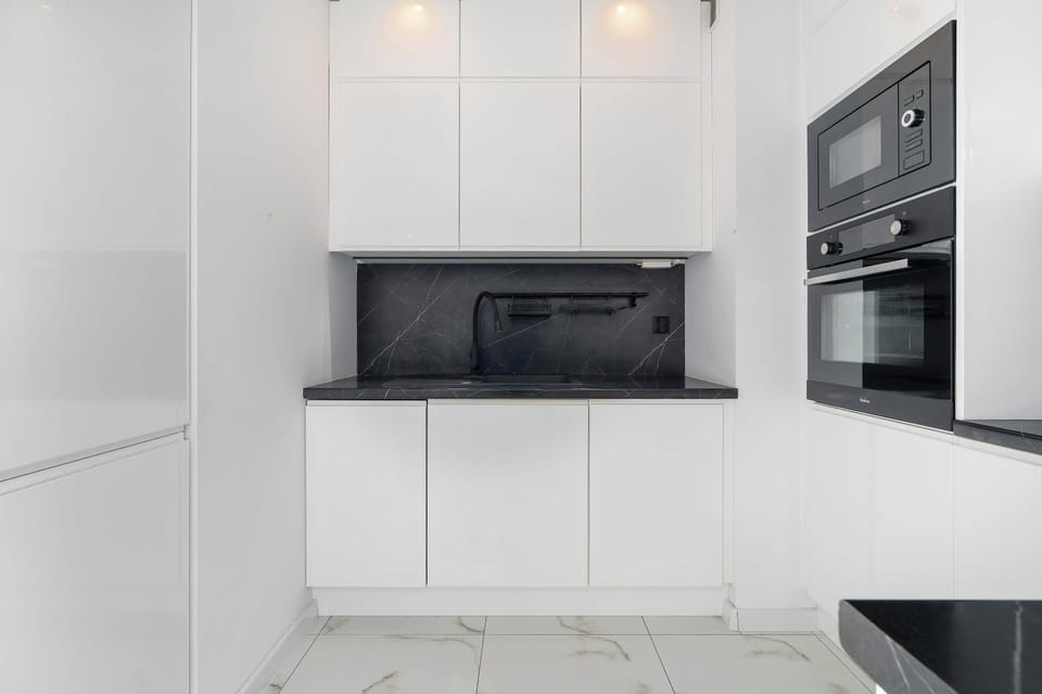 The modern kitchenette features a built-in microwave and oven integrated into seamless white cabinetry, contrasted by a black backsplaslı.