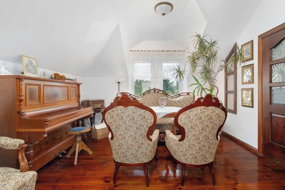 A dining space arranged with upholstered chairs and a polished wooden table beside a piano. Natural light enters through multiple windows under the vaulted ceiling.