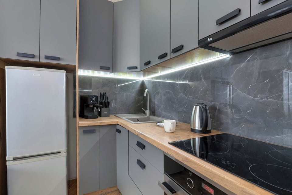 A modern compact kitchen boasts sleek grey cabinetry, a built-in oven, an induction cooktop, and a freestanding silver refrigerator against a dark backsplash.