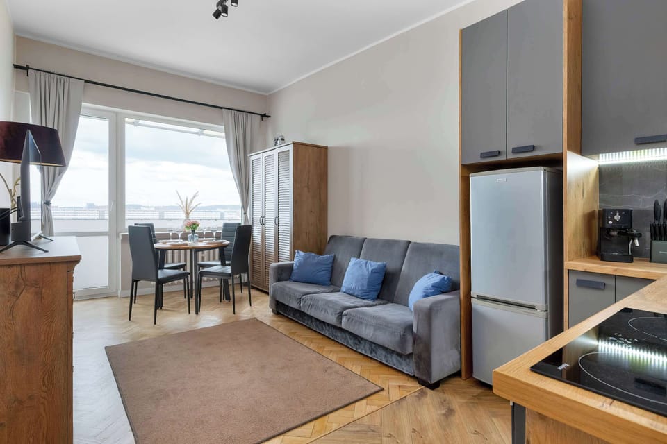 An open-concept living space seamlessly transitions from the kitchen area to a living room setup with a grey sofa, a wooden credenza, and a dining table near the window.