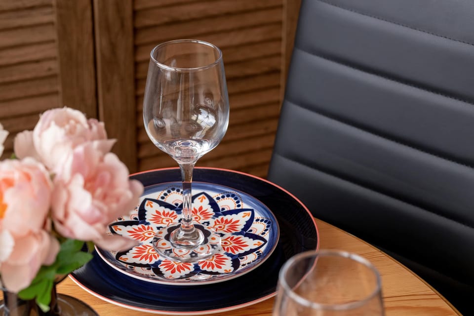 A detailed close-up captures a beautifully arranged place setting on a wooden table, featuring an intricate patterned plate, a wine glass, and a delicate bouquet of pink flowers.