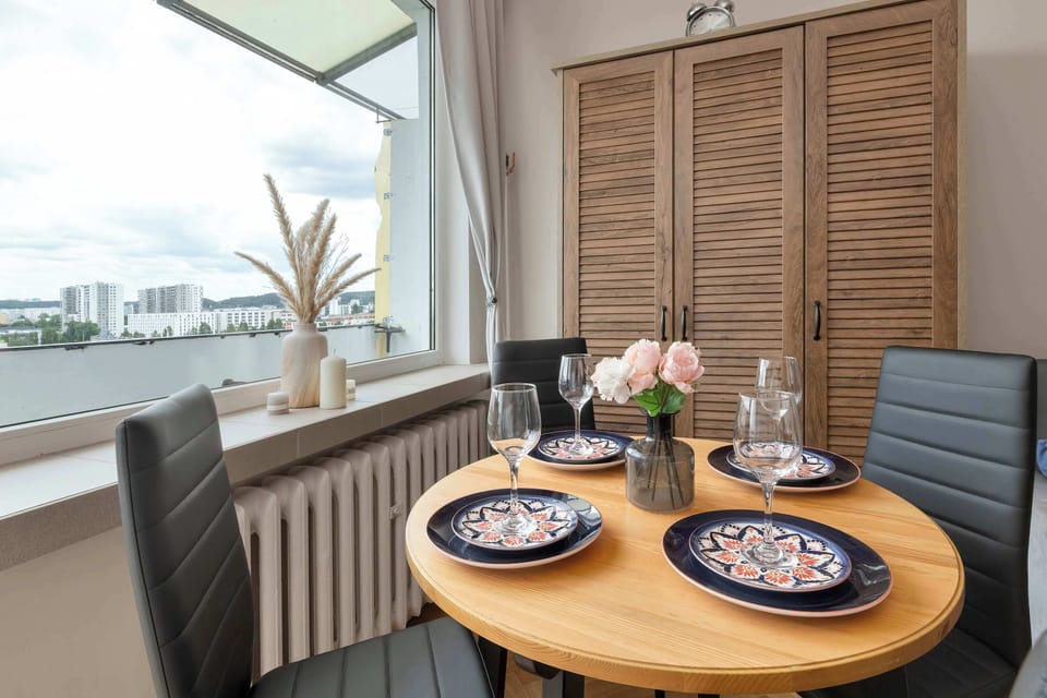 A charming dining nook by a wide window offers a view of the outdoors, centered around a round wooden table decorated with a vase of dried pampas grass.