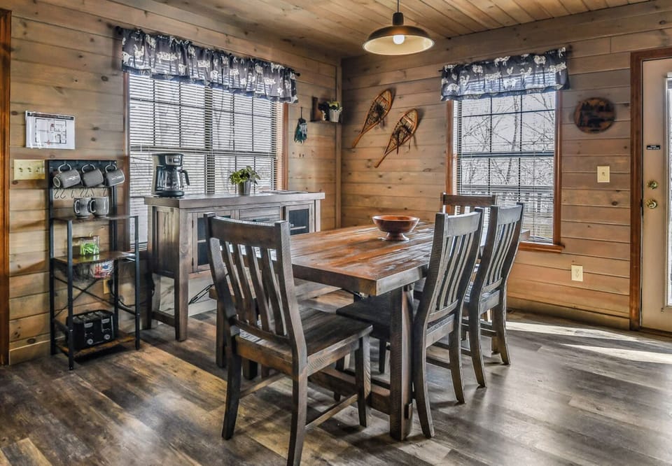 Your rustic dining space invites shared meals under warm pendant lighting, with handcrafted wood furnishings and charming snowshoe decor. Natural light streams through curtained windows framing peaceful winter views.