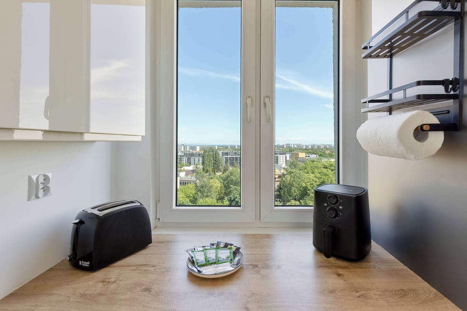 A bright window in the kitchen offers an expansive view of green treetops under a clear blue sky. A black toaster and an electric kettle sit on the wooden countertop below.