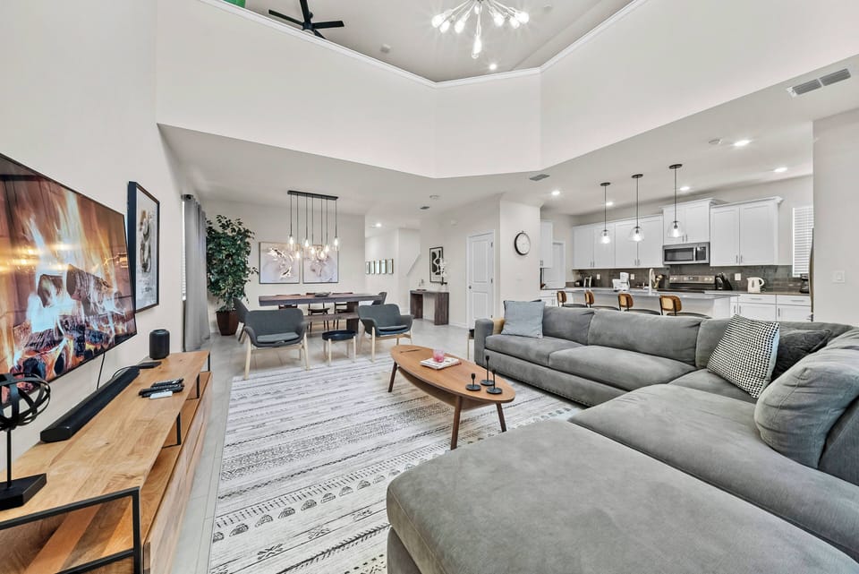 Welcome to a bright and spacious living area featuring high ceilings, a sleek gray sectional, and warm wood accents. The open layout flows into a stylish dining area, ideal for sharing moments after your day's adventures.