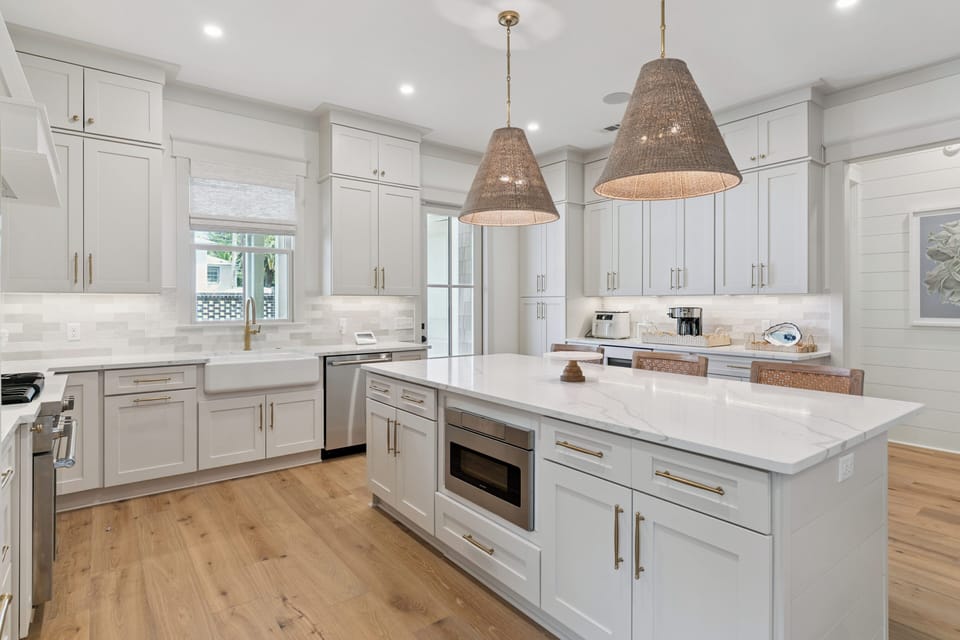 Picture yourself preparing meals in this bright, elegant kitchen with gleaming white cabinetry, spacious marble island, and warm coastal lighting—perfect for gathering and creating memories.