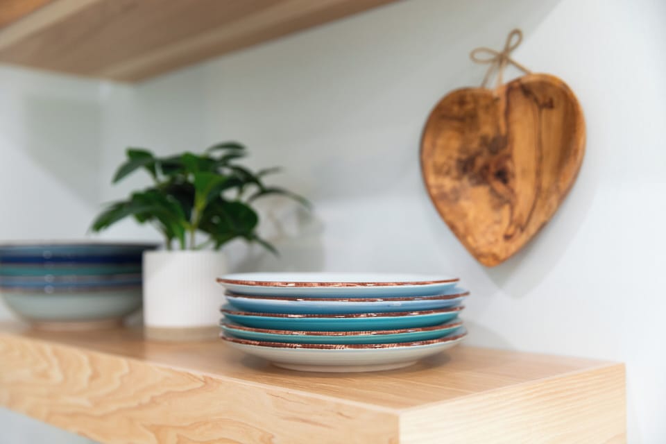 A close-up view of the natural wood floating shelves showcases a stack of glazed turquoise side plates alongside a heart-shaped wooden bowl suspended by a twine bow.