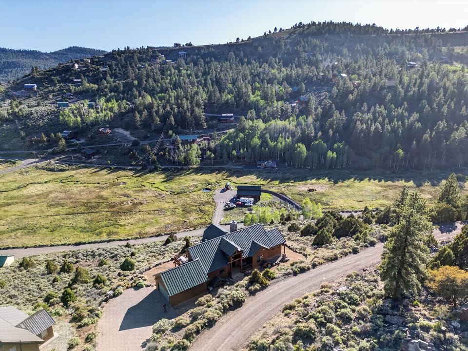 Aerial view of the vacation rental property nestled in a scenic mountain valley, surrounded by lush pine forests and rolling meadows with neighboring homes dotting the hillsides.
