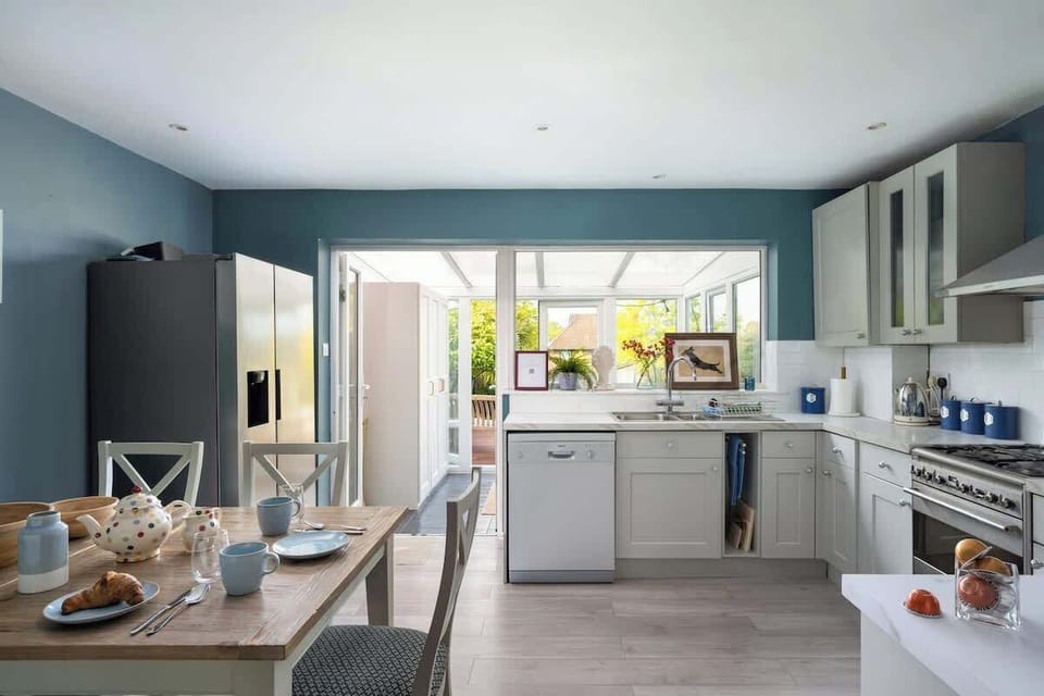 Contemporary Heart
A bright, open kitchen in image_ddfb3b.png pairing muted teal walls with classic white cabinetry. An integrated dining area flows directly into a sunlit conservatory, blending functional design with breezy charm.