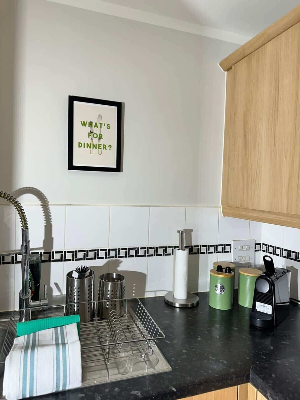 The well-appointed kitchen counter features a stylish mixer tap, dark granite worktop, and all essential cooking tools. A cheeky 'What's for dinner?' print on the wall sets the tone for fun, relaxed evenings cooking in.