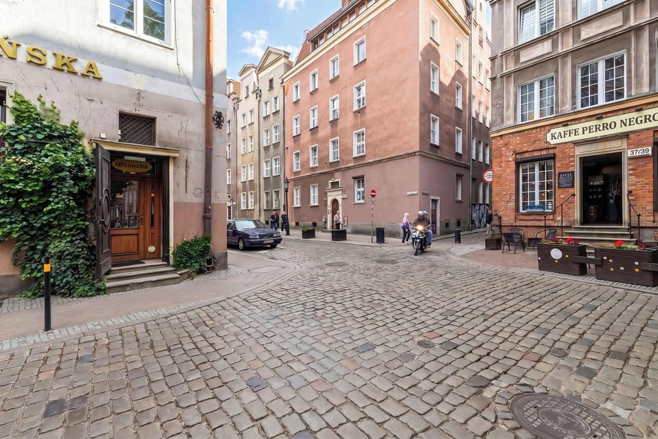 A charming cobbled courtyard bordered by historic buildings with storefronts and residential entrances. Outdoor greenery softens the urban setting.