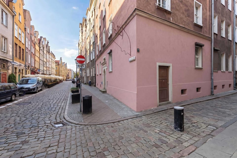 A cobblestone street corner lined with colorful historic buildings. The pink façade at the intersection stands out against the surrounding architecture.