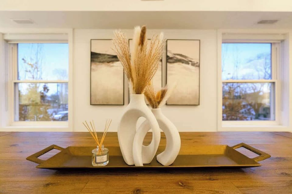 Elegant wooden tray centerpiece with sculptural vases filled with decorative dried grasses and a fragrant reed diffuser, set against large windows and artistic wall paintings.
