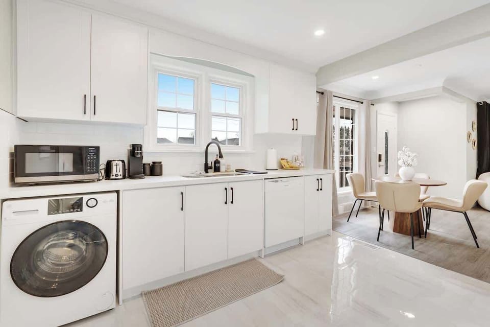Bright, spacious kitchen featuring modern white cabinetry, front-load washer, microwave, coffee maker, toaster, blender, dishwasher, double windows, and an elegant dining area with stylish chairs and table.