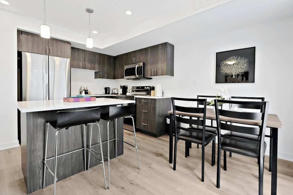 Modern, spacious kitchen with sleek stainless steel appliances, elegant island with bar stools, cozy dining table, stylish cabinetry, and tasteful wall art for comfort.