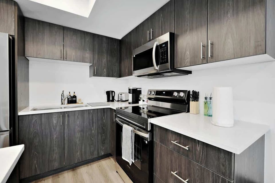 Sleek, modern kitchen with stainless steel appliances, spacious countertops, ample dark wood cabinets, coffee maker, knife set, paper towel holder, and stylish finishes.