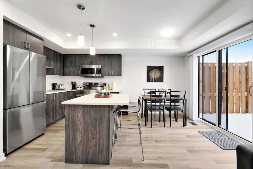 Bright modern kitchen with large island seating, stainless steel fridge, microwave, stove, toaster, coffee maker, dining table, chairs, and sliding glass doors to patio.
