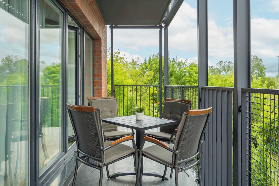The balcony dining area captures the square outdoor table and four woven mesh chairs up close. Thick green treetops are visible beyond the safety railing, offering a pleasant view.
