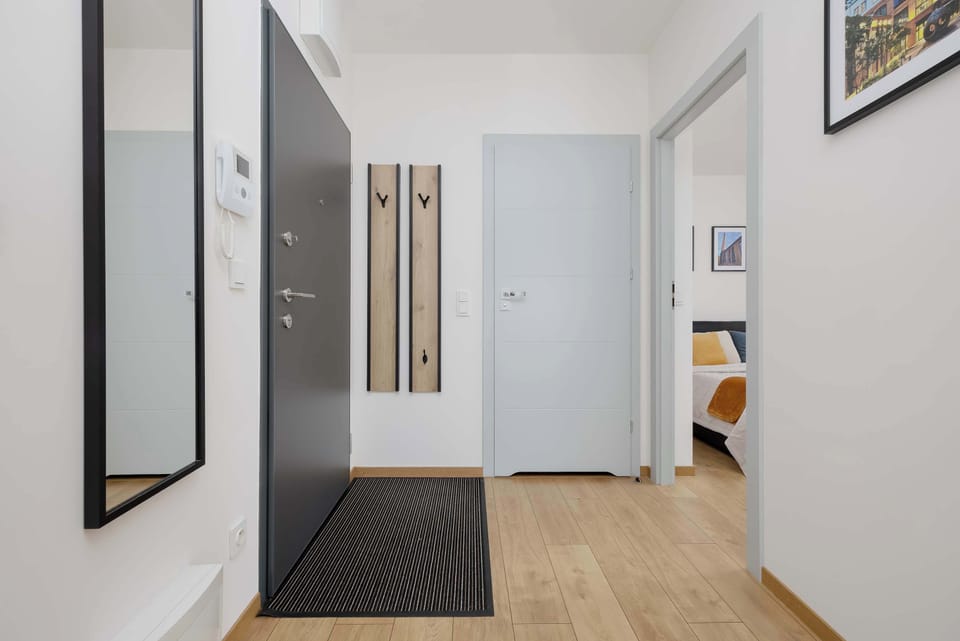 A minimalist hallway features a full-length wall mirror and a dark gray entry door with integrated coat hooks. A simple black-and-white patterned rug lines the wood-plank floor.