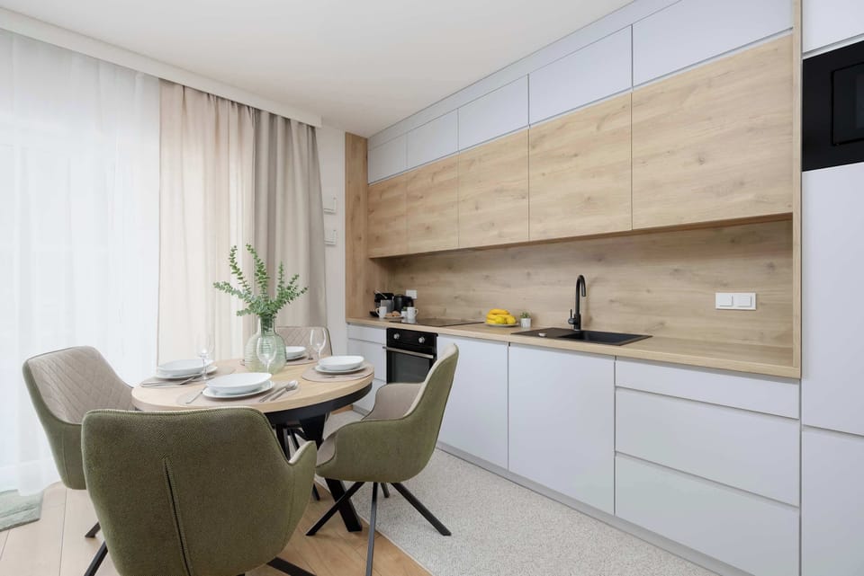 The dining area is set with a round wooden table and plush, sage green upholstered chairs. In the background, a modern kitchen with light wood upper cabinets and white lower units lines the wall.