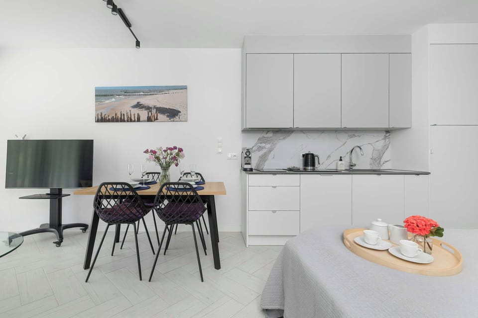 An open perspective connects the dining table to the kitchen area, showing a flat-screen TV on a standalone mount to the left. A decorative tray with hospitality items rests on a bed frame in the foreground corner.