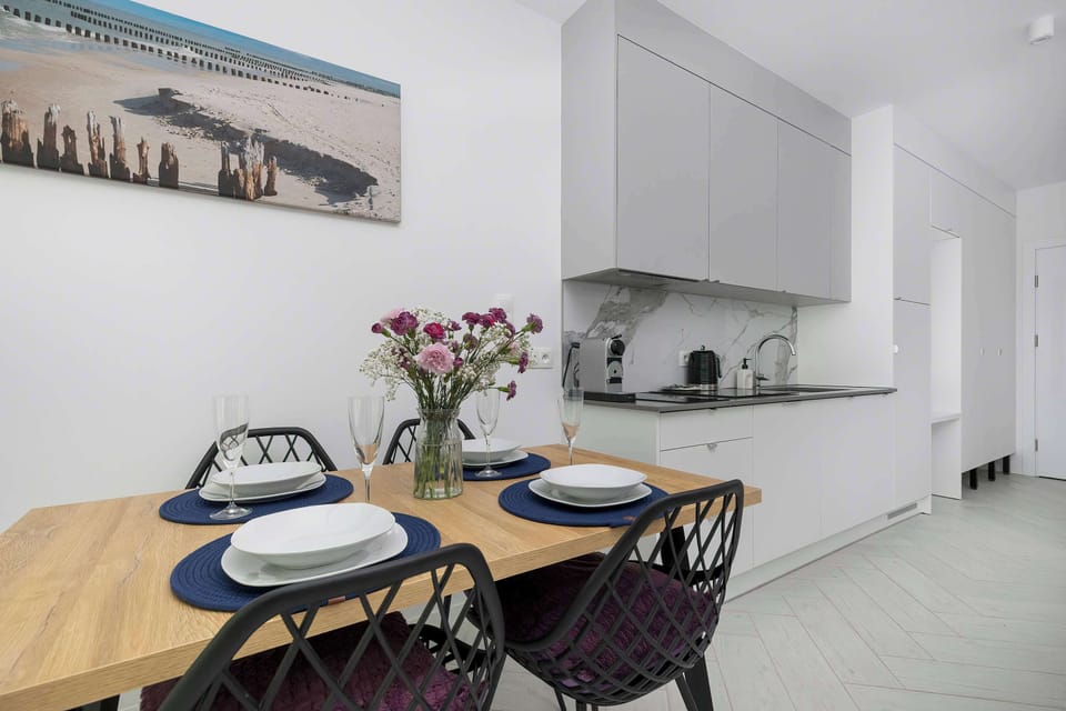 A dining area with a wooden table is positioned near a compact gray kitchenette setup equipped with modern appliances. A colorful framed artwork depicting a coastline hangs on the wall, adding a touch of personality to the space.