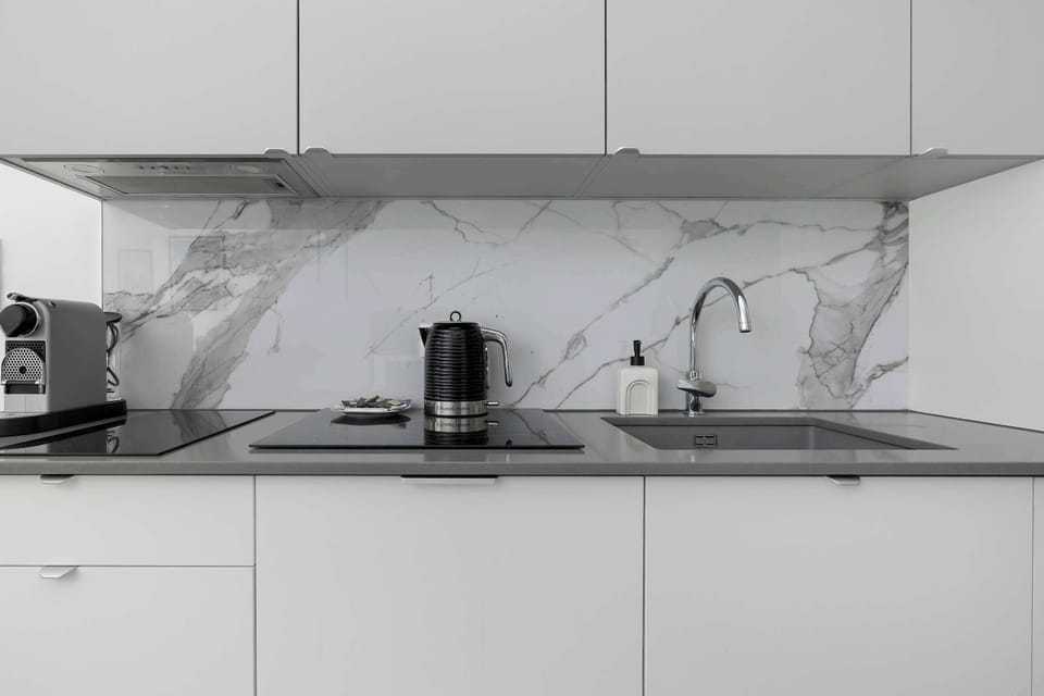 A sleek, minimalist kitchenette features handleless white cabinetry beneath a striking white marble backsplash with grey veining. A black espresso maker and electric kettle sit neatly on the clean countertop surface.