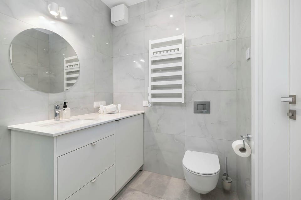 A bright bathroom setup features a floating white vanity with a round mirror and an illuminated chrome towel radiator on the wall. A modern wall-mounted toilet is positioned in the corner against the light gray tiling.